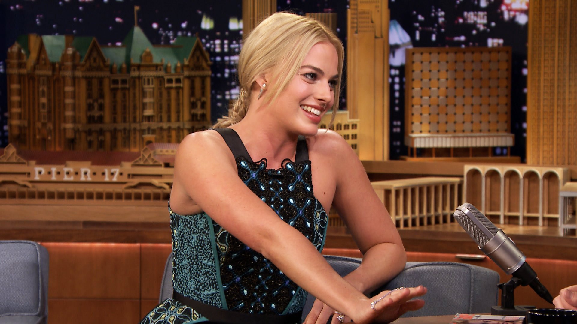HD desktop wallpaper featuring celebrity Margot Robbie smiling and engaging in conversation on a late-night talk show set.