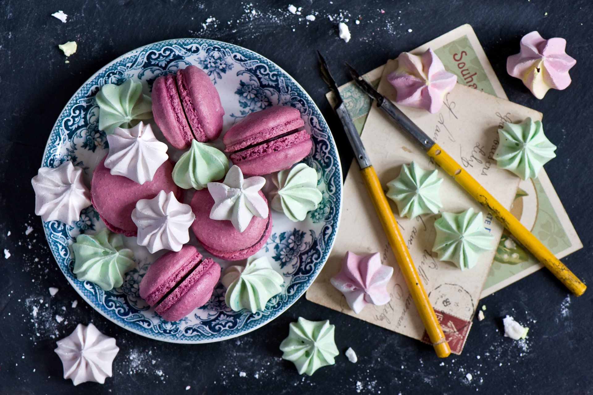 A HD desktop wallpaper featuring colorful meringue kisses and pink macarons arranged on a decorative plate alongside paintbrushes and vintage paper.