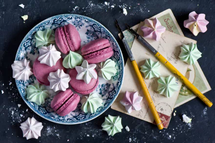 A HD desktop wallpaper featuring colorful meringue kisses and pink macarons arranged on a decorative plate alongside paintbrushes and vintage paper.