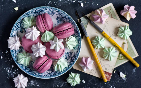 A HD desktop wallpaper featuring colorful meringue kisses and pink macarons arranged on a decorative plate alongside paintbrushes and vintage paper.