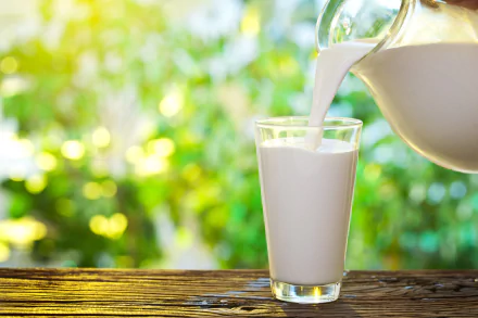 A high-definition desktop wallpaper featuring milk being poured from a glass jug into a glass, set against a vibrant, green, sunlit background.