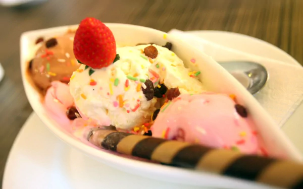 A vibrant HD desktop wallpaper featuring a colorful banana split with scoops of ice cream, whipped cream, sprinkles, a strawberry, and a chocolate wafer on a white plate.