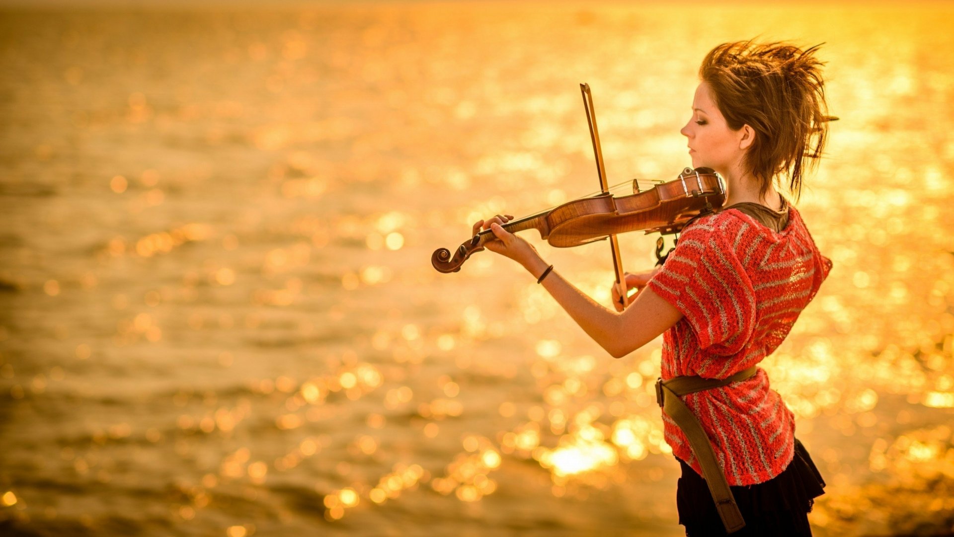 Lindsey Stirling passionately plays the violin by a shimmering golden water backdrop in this HD PC desktop wallpaper showcasing music and artistry.