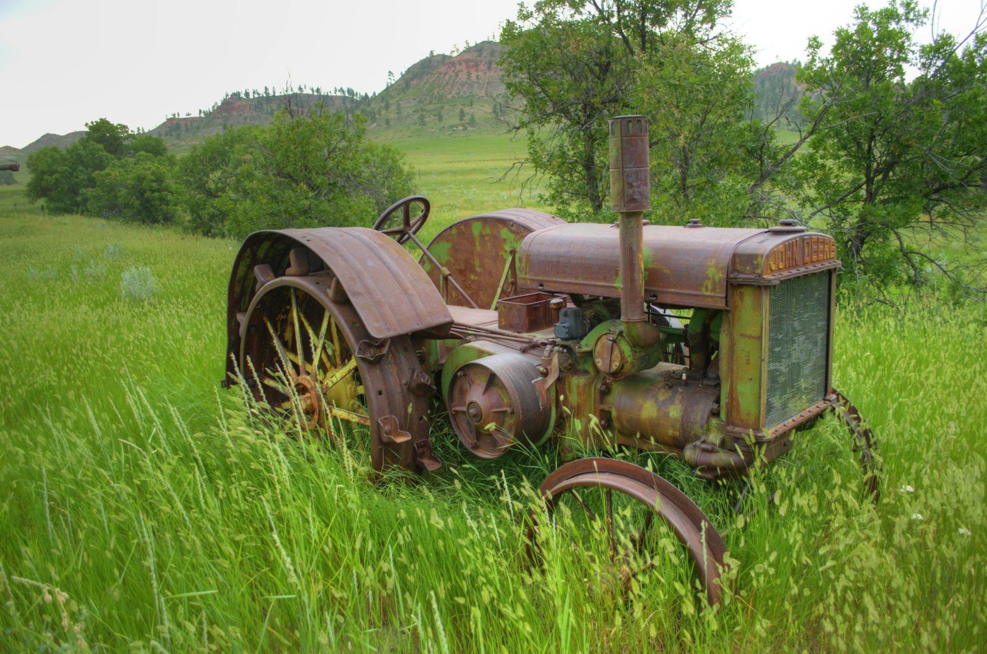 Rusty old John Deere tractor sits abandoned in tall grass with a scenic hill and trees in the background, captured as a vibrant HD desktop wallpaper.