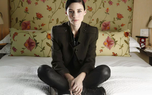 HD PC desktop wallpaper: a celebrity woman with dark hair and a dark jacket sitting cross-legged on a bed with a floral headboard.