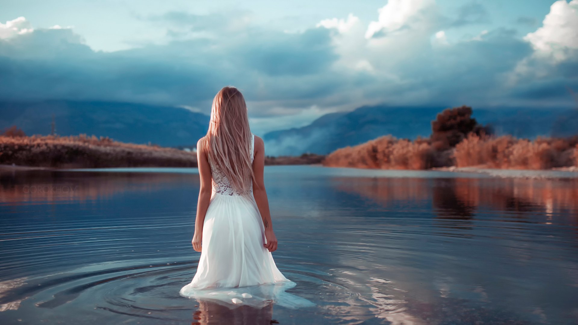 A blonde woman in a white dress stands in shallow water outdoors, facing serene mountains under a cloudy sky. HD desktop wallpaper and background.