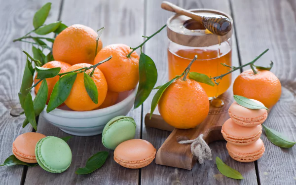 HD desktop wallpaper featuring vibrant mandarins, a jar of honey with a dipper, and colorful macarons arranged on a rustic wooden surface.