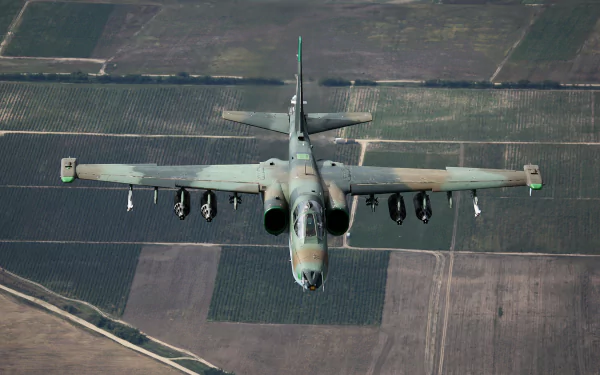 A Sukhoi Su-25 military aircraft soars above a vast landscape, showcasing its unique camouflage and weaponry, set against a backdrop of verdant fields. HD desktop wallpaper.