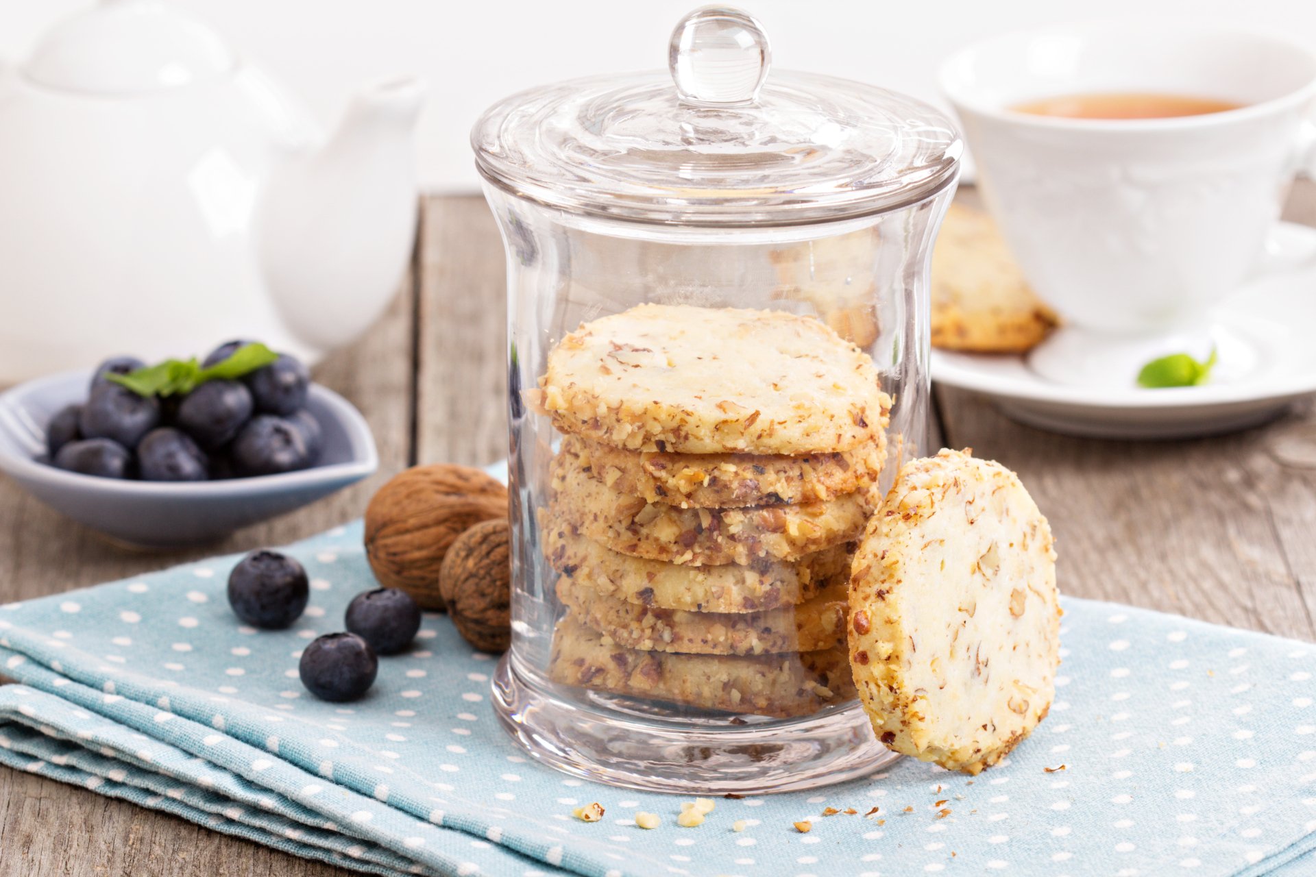 Clear glass jar filled with walnut cookies on a blue polka dot cloth, accompanied by fresh blueberries, tea in a white cup, and a white teapot in a bright, 4K Ultra HD setting.