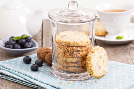 Clear glass jar filled with walnut cookies on a blue polka dot cloth, accompanied by fresh blueberries, tea in a white cup, and a white teapot in a bright, 4K Ultra HD setting.