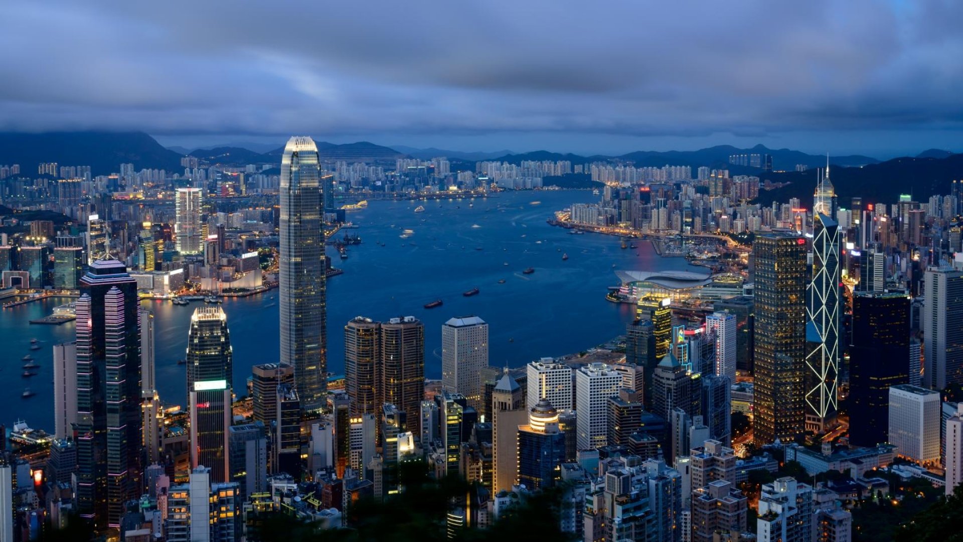A vibrant 4K Ultra HD man-made cityscape of Hong Kong at dusk, showcasing illuminated skyscrapers and boats on the harbor against a cloudy sky.