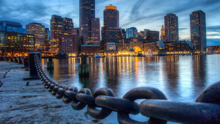 A stunning HDR view of the Boston skyline at dusk, showcasing sparkling city lights reflected in the water, framed by a foreground chain along the shore. 4K Ultra HD wallpaper.