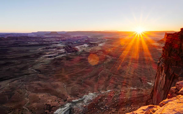 4K Ultra HD desktop wallpaper showing sunbeams bursting over a vast canyon at sunrise, warm light illuminating red cliffs, layered rock formations and a winding valley below.