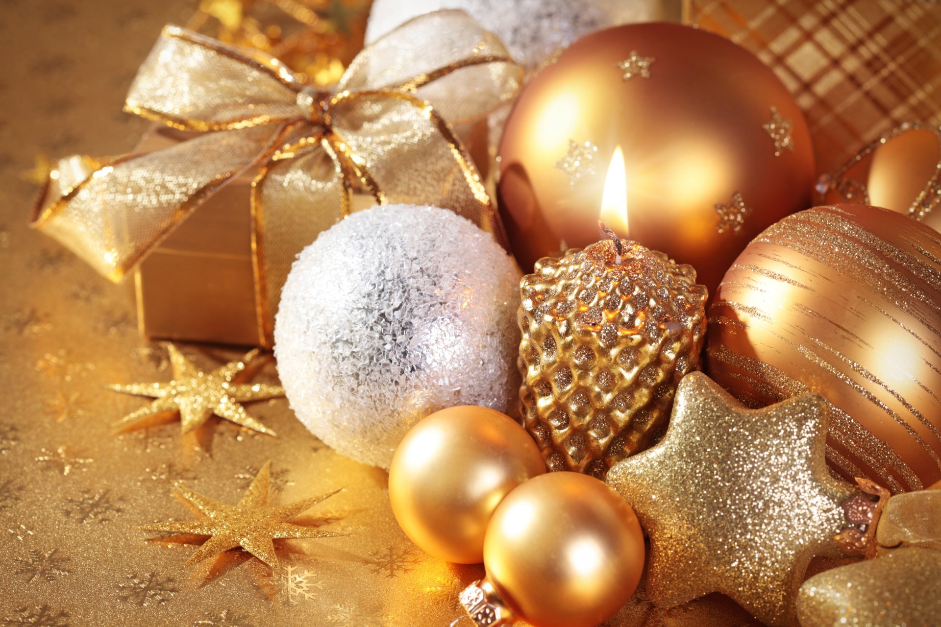 A festive arrangement featuring a lit candle surrounded by gold Christmas ornaments, sparkling stars, and wrapped presents, creating a warm holiday ambiance.