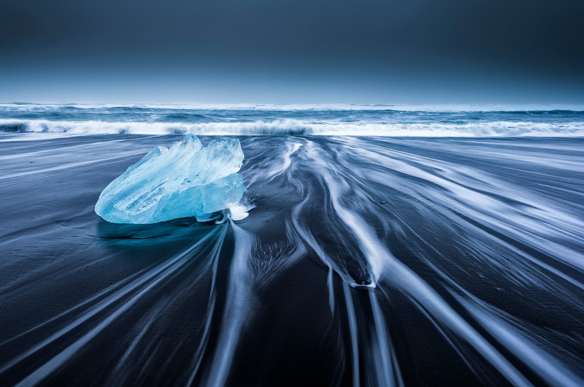 HD desktop wallpaper of a serene beach with dark sand, gentle ocean waves, a piece of ice on shore, and a clear horizon under a deep blue sky.