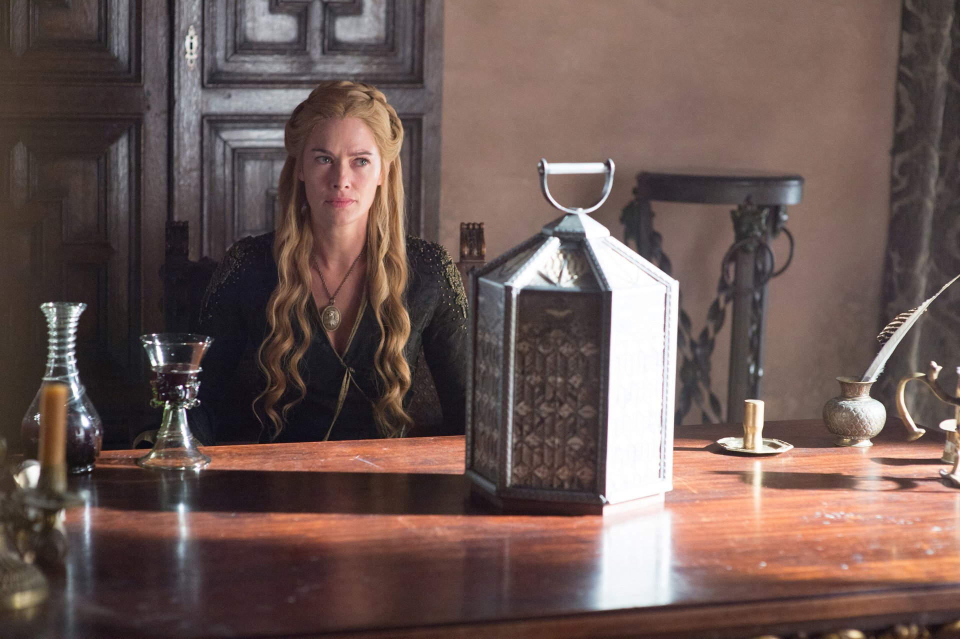 Cersei Lannister, portrayed by Lena Headey in the TV show Game of Thrones, sits at a wooden table in a dimly lit room, captured in HD for a PC desktop wallpaper.