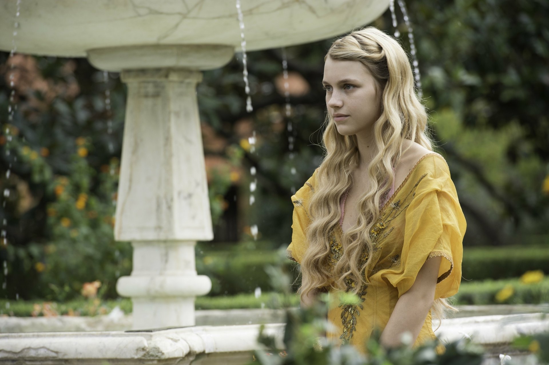 Nell Tiger Free as Myrcella Baratheon from Game of Thrones stands near a fountain in a lush garden, captured in a 4K Ultra HD TV show wallpaper.