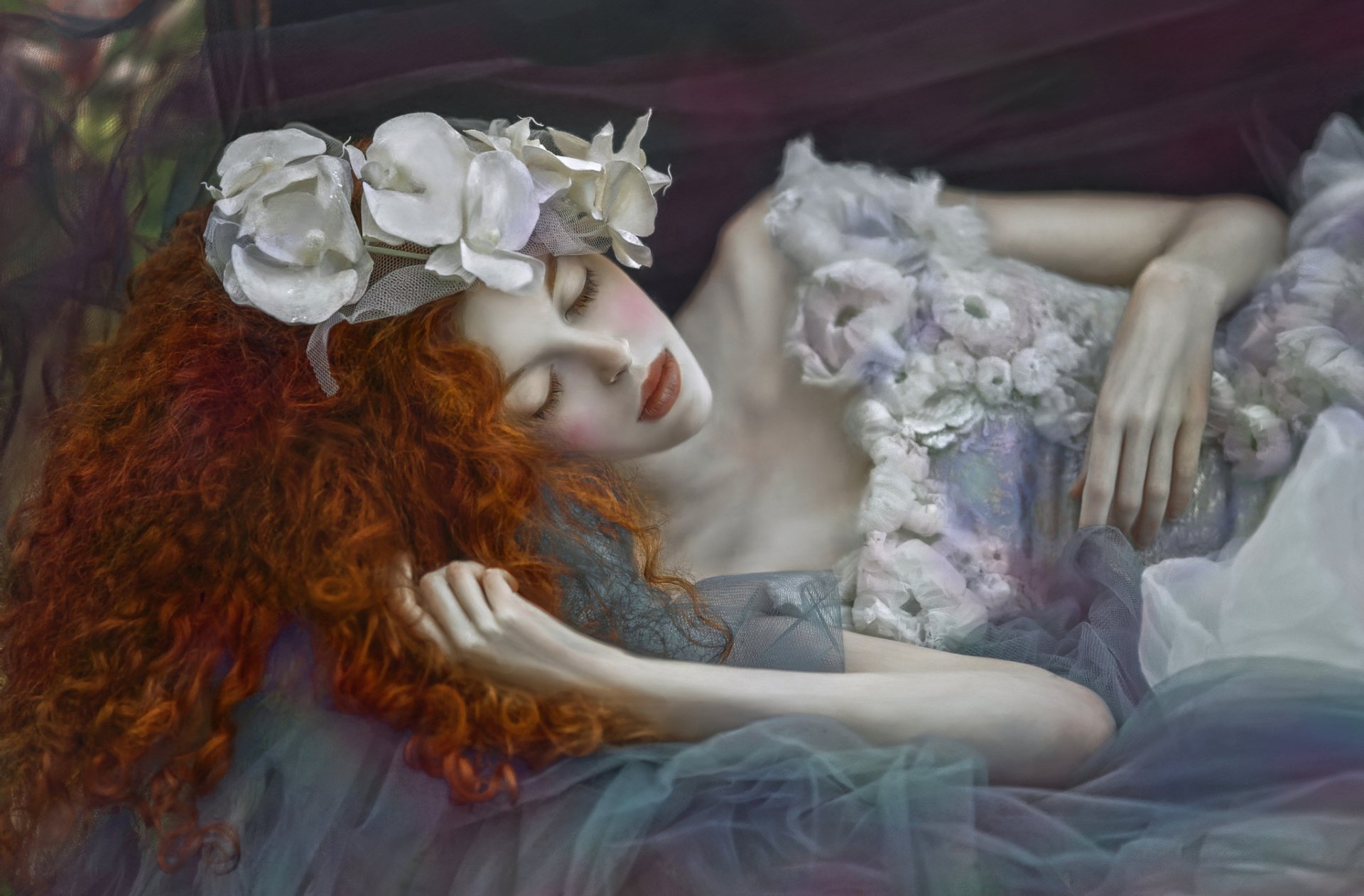HD PC desktop wallpaper and background: dreamy mood portrait of a redhead woman with long flowing hair, wearing a floral crown, reclining amid soft pastel fabrics.