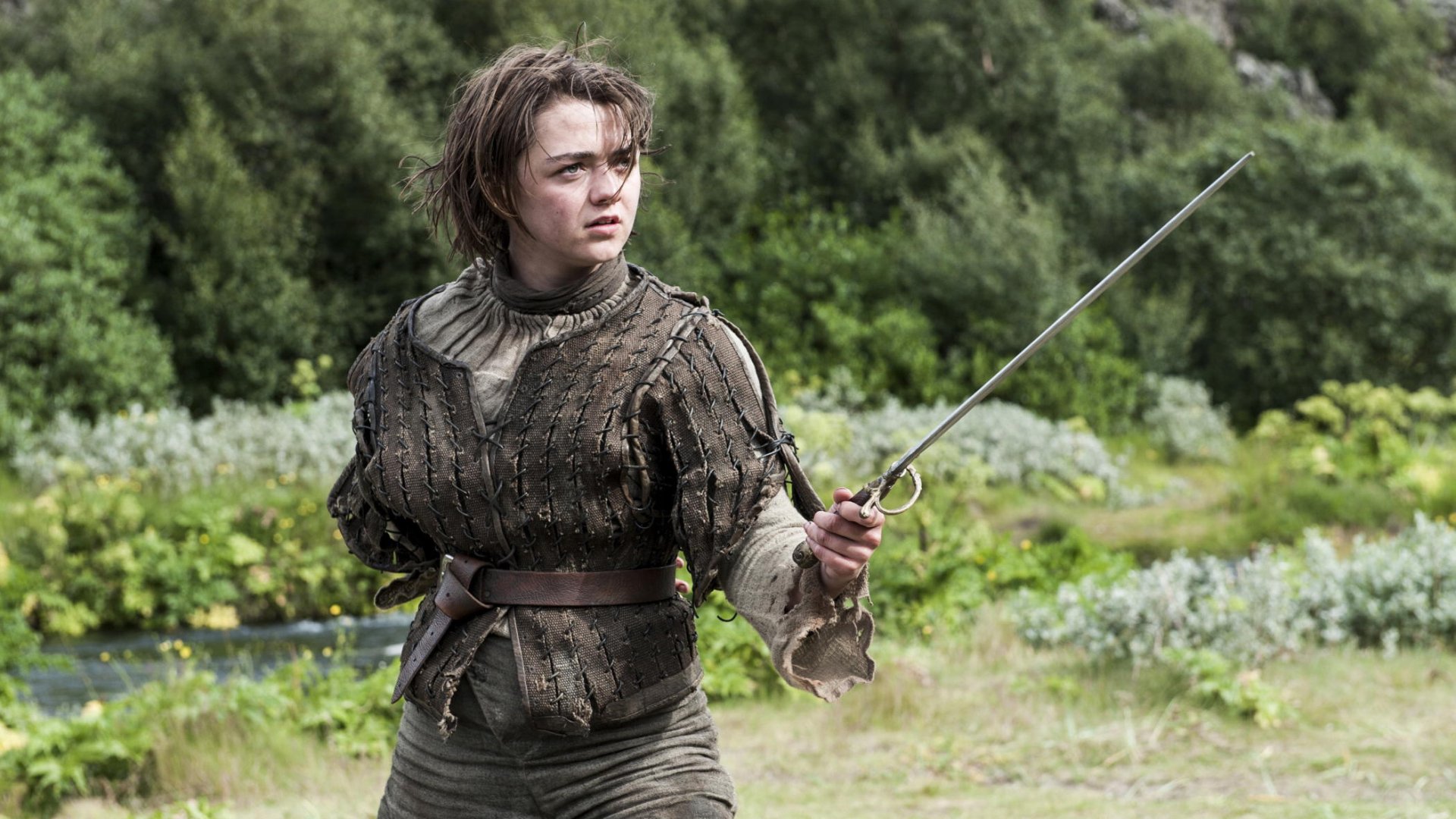 Maisie Williams as Arya Stark poses with a sword in this HD Game of Thrones desktop wallpaper, showcasing her fierce character in a lush outdoor setting.
