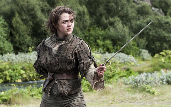 Maisie Williams as Arya Stark poses with a sword in this HD Game of Thrones desktop wallpaper, showcasing her fierce character in a lush outdoor setting.