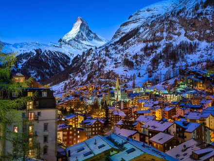 Snow-covered village nestled in the Swiss Alps at dusk, with warm lights glowing under the iconic Matterhorn peak in winter.