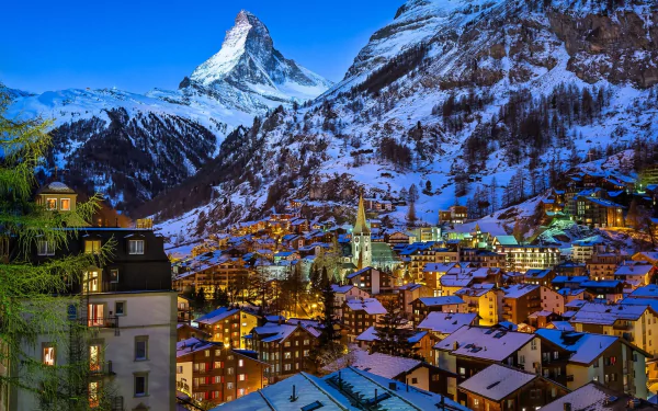 Snow-covered village nestled in the Swiss Alps at dusk, with warm lights glowing under the iconic Matterhorn peak in winter.