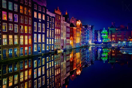 A vibrant night scene of Amsterdam's iconic colorful buildings along the canal, beautifully reflected in the water, creating a stunning HD desktop wallpaper.