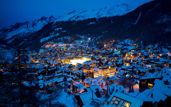 Nighttime cityscape of snowy Zermatt in the Swiss Alps, with glowing lights illuminating the winter town nestled in a valley under clear blue skies.