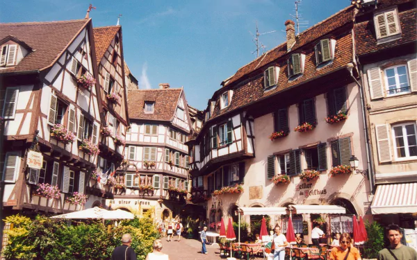 HD desktop wallpaper showcasing the charming man-made architecture of Colmar with timber-framed buildings and lively street cafes under a clear blue sky.
