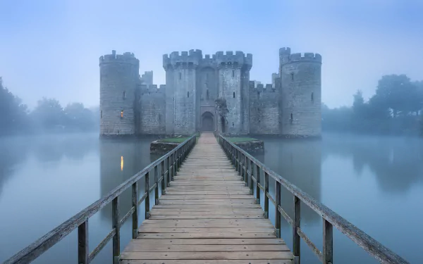 Fog envelops Bodiam Castle reflected in the calm water, with a wooden bridge leading to its medieval stone walls in this HD PC desktop wallpaper.