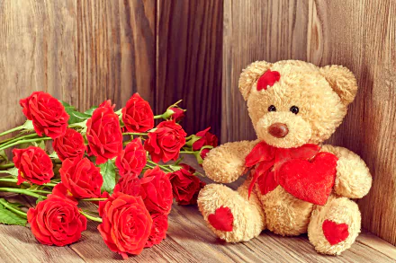 A HD desktop wallpaper featuring a cute teddy bear with red heart accents, sitting beside a bunch of vibrant red roses, set against a wooden background, evoking a romantic Valentine's Day ambiance.