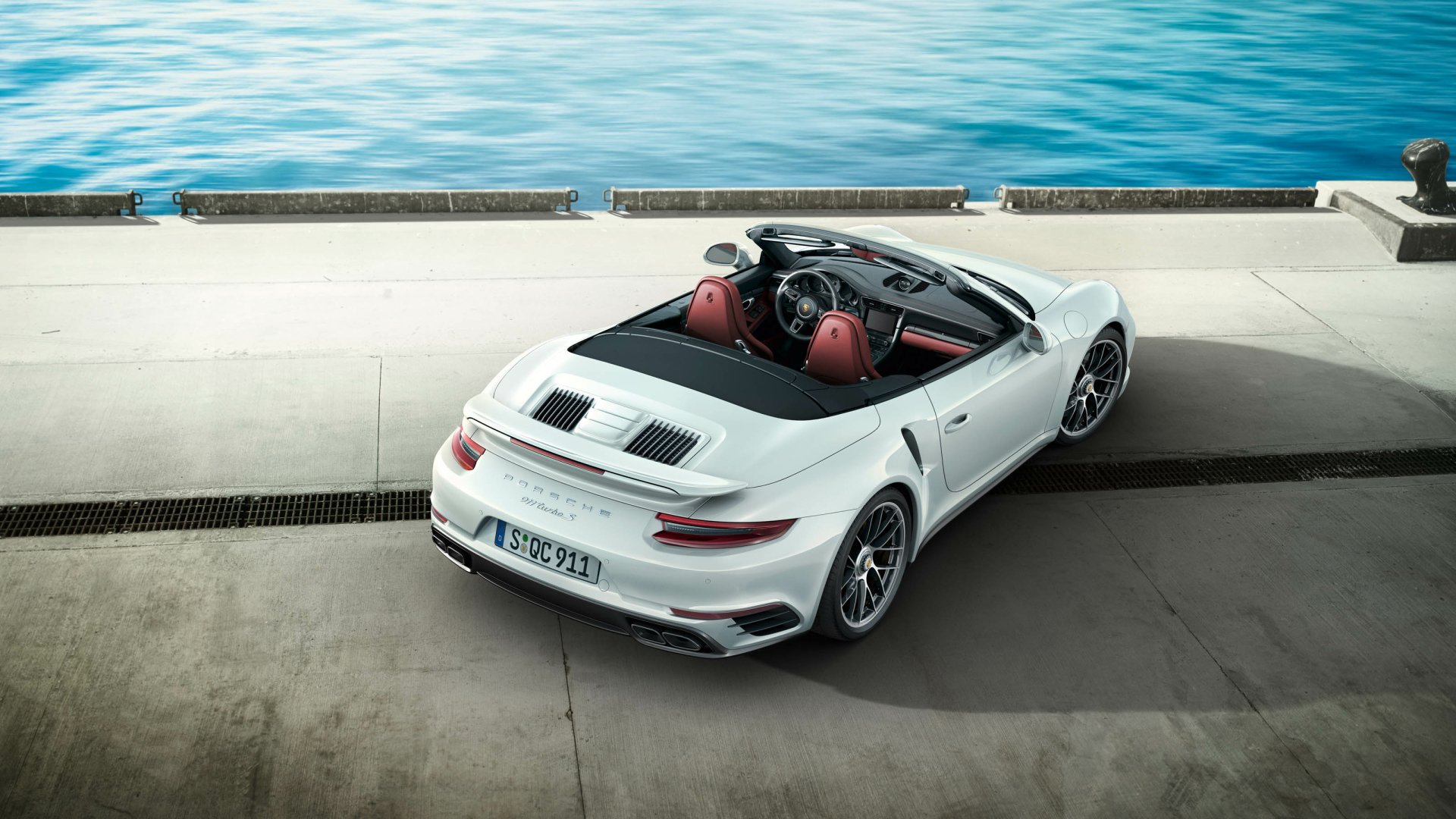 Silver Porsche 911 Turbo convertible parked on a waterfront pier with a calm blue sea in the background, captured in HD for a desktop wallpaper.