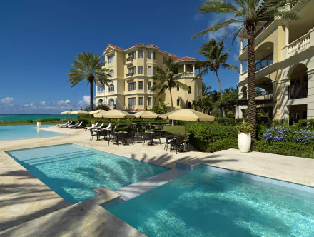 4K Ultra HD view of a Turks and Caicos hotel with palm trees, ocean in the background, and a man-made pool surrounded by elegant architecture and outdoor seating.