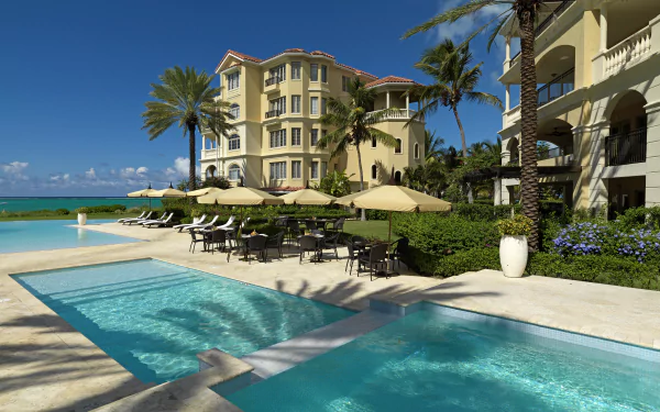 4K Ultra HD view of a Turks and Caicos hotel with palm trees, ocean in the background, and a man-made pool surrounded by elegant architecture and outdoor seating.