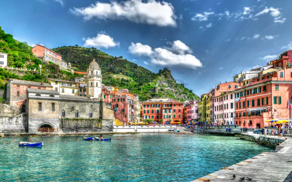 Vibrant 4K HDR view of Manarola, Italy, showcasing colorful buildings, a coastal dome, boats on clear water, and lush mountains under a bright sky.