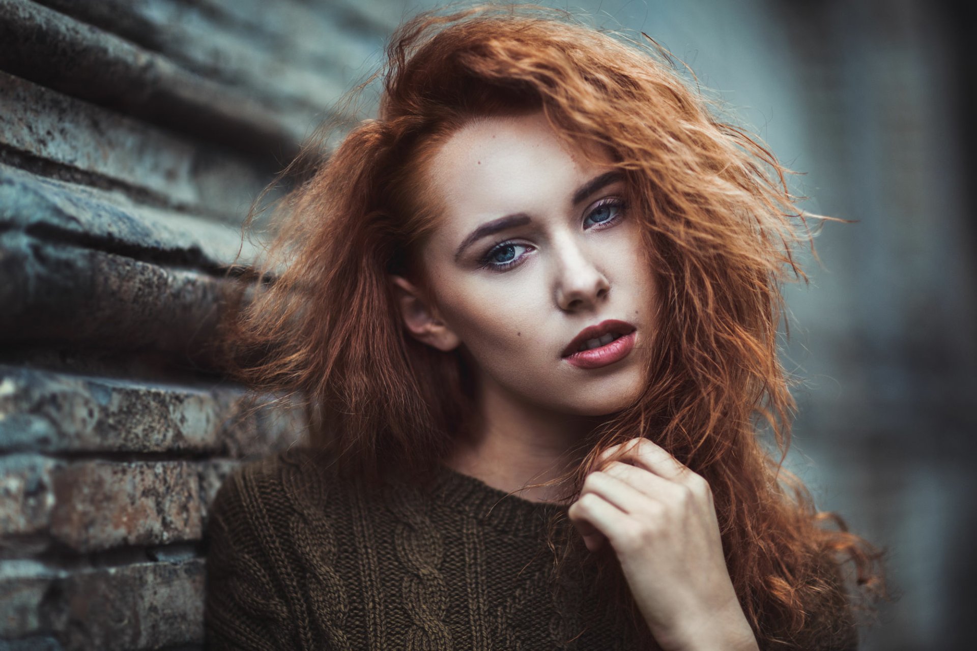 HD desktop wallpaper of a redhead woman model with striking blue eyes and wavy hair, posed against a textured brick wall background.