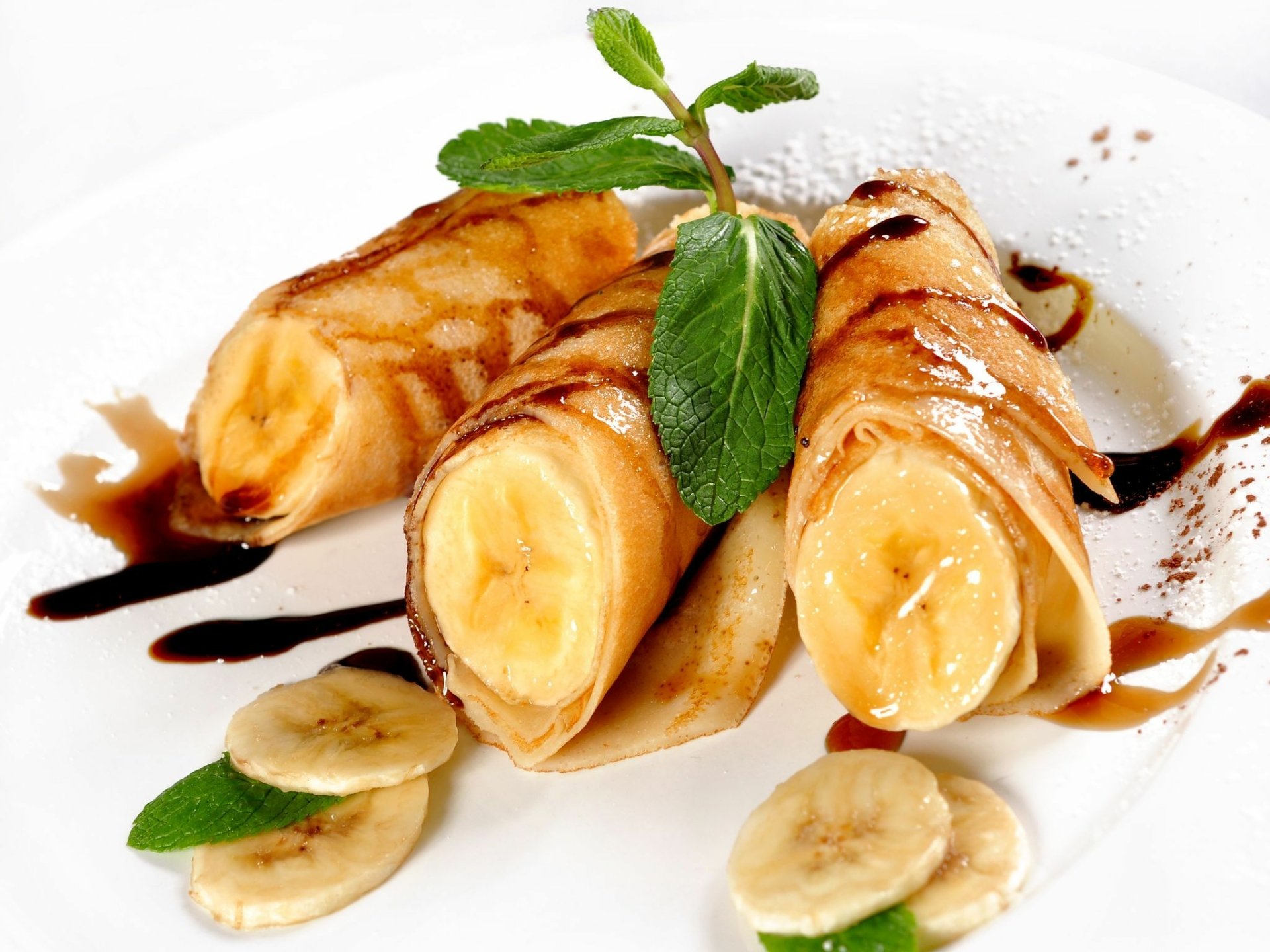 HD desktop wallpaper of banana-filled crêpes drizzled with chocolate sauce, garnished with fresh mint leaves and banana slices on a white plate.