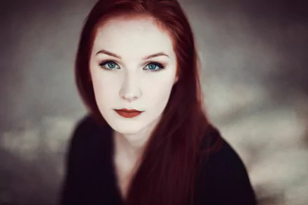 A striking redhead with bold lipstick and captivating blue eyes gazes directly at the viewer, set against a soft bokeh background, creating an elegant and artistic portrait.