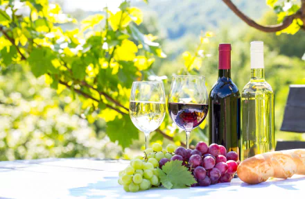 4K Ultra HD still life desktop wallpaper featuring glass bottles of wine, two wine glasses, and clusters of green and red grapes on an outdoor table with greenery in the background.