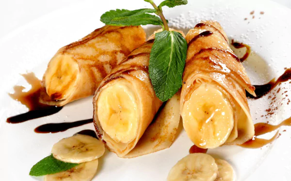 HD desktop wallpaper of banana-filled crêpes drizzled with chocolate sauce, garnished with fresh mint leaves and banana slices on a white plate.