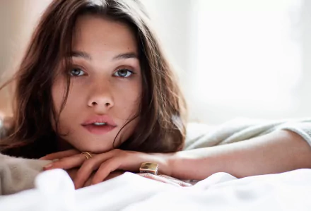 A close-up portrait of brunette French actress and model Astrid Bergès-Frisbey, gazing thoughtfully while resting on a soft surface. The image features natural lighting and soft tones.