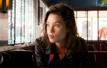 A brunette woman with striking green eyes, dressed in a stylish leather jacket, gazes thoughtfully while seated in a trendy café setting. A captivating image of French actress Astrid Bergès-Frisbey.