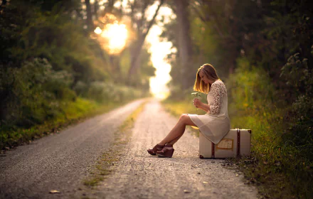 A blonde woman sits on a suitcase in the middle of a sunny, deserted road, surrounded by lush greenery, captured in a calm, reflective mood. The setting sun adds warmth to the outdoor HD wallpaper background.