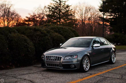 HD desktop wallpaper featuring a sleek silver Audi S4 parked on an asphalt road with trees and a sunset sky in the background.