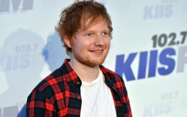 Smiling red-haired man in a red-and-black plaid shirt standing before a radio-station music backdrop — 2K Quad HD PC desktop wallpaper.