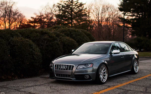 HD desktop wallpaper featuring a sleek silver Audi S4 parked on an asphalt road with trees and a sunset sky in the background.