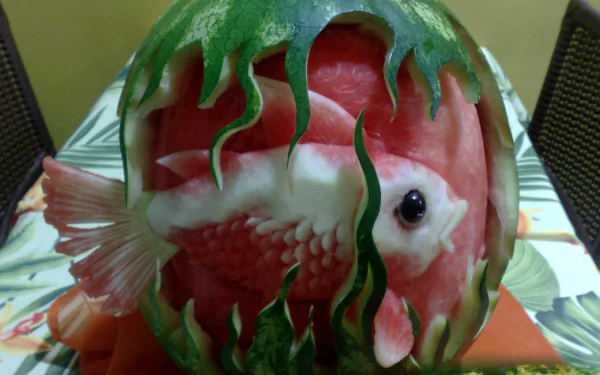 Watermelon carving of a lifelike fish with seaweed details, vibrant food art photographed as a 2K Quad HD PC desktop wallpaper and background.