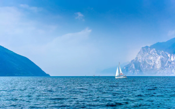 A serene blue ocean stretches to the horizon, with a sailboat gliding peacefully on the water, framed by majestic mountains in the distance. 4K Ultra HD wallpaper.