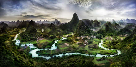 HD desktop wallpaper showcasing a breathtaking landscape of the Li River meandering through a village, fields, and lush forests in the Nanling Mountains, Guanxi Zhuang, China, under a partly cloudy sky.