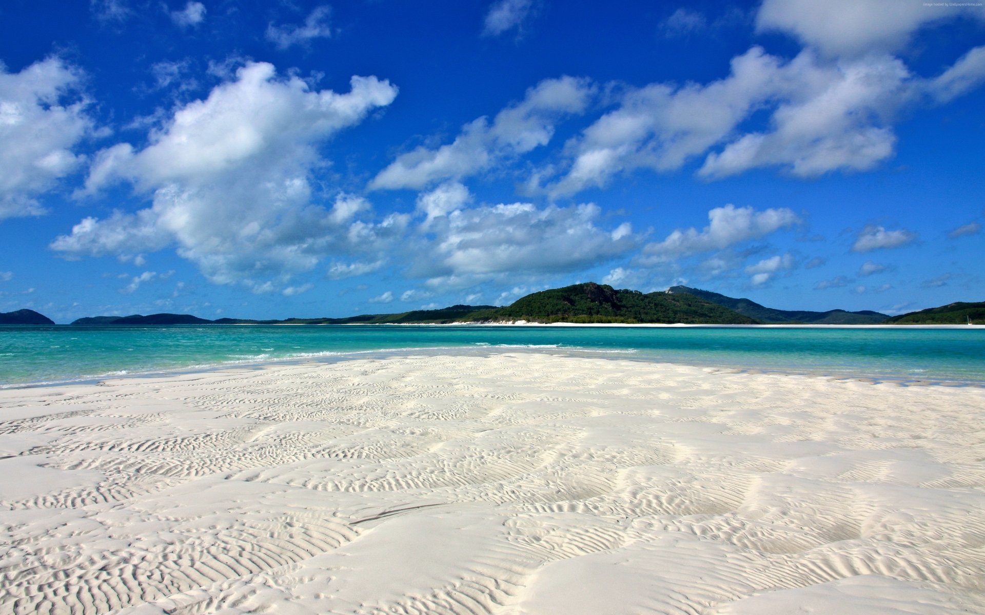 Turquoise tropical ocean meets white sandy beach under a bright blue sky with clouds along the coastline of Australia in stunning 4K Ultra HD resolution.